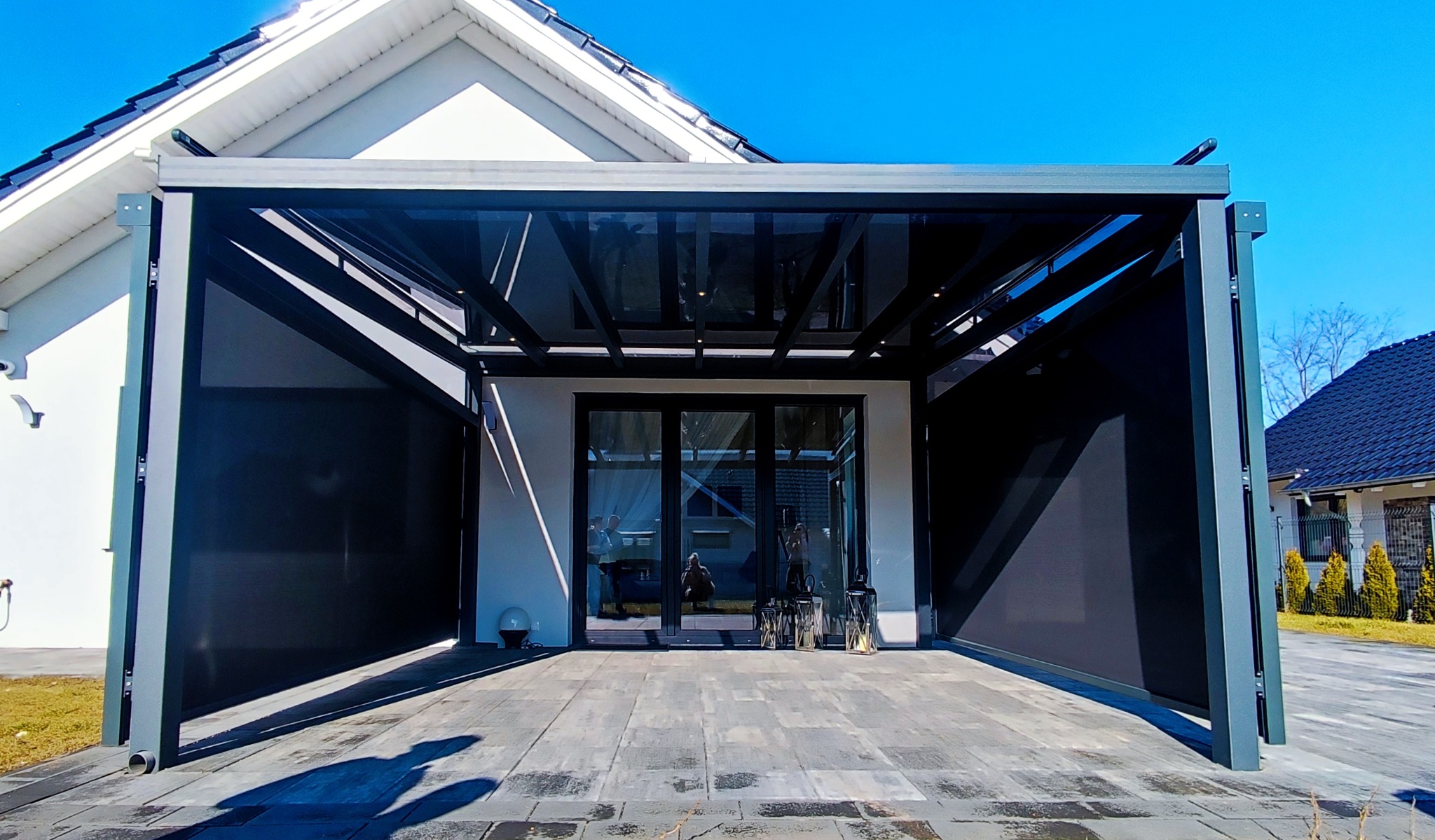 Nowoczesna pergola z metalową konstrukcją i roletami zaciemniającymi, przylegająca do domu z dużym oknem tarasowym i szarą kostką brukową.