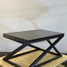 ArtLoftHome - Stolik kawowy z ciemnego drewna i czarną, spawana metalową podstawą w kształcie litery X, na podłożu z płyty OSB, na tle jasnej ściany.