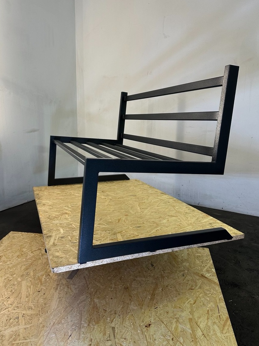 Metalowy stelaż sofy w stylu industrialnym, czarne profile na tle surowej płyty OSB. Minimalistyczny design, spawane łączenia, widoczne niedoskonałości powierzchni.