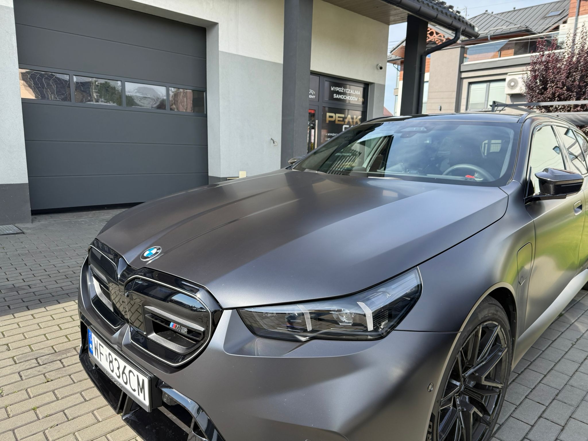 Nowoczesny, matowy, grafitowy samochód BMW kombi z widocznymi detalami, zaparkowany przed budynkiem wypożyczalni samochodów Peak Rent.