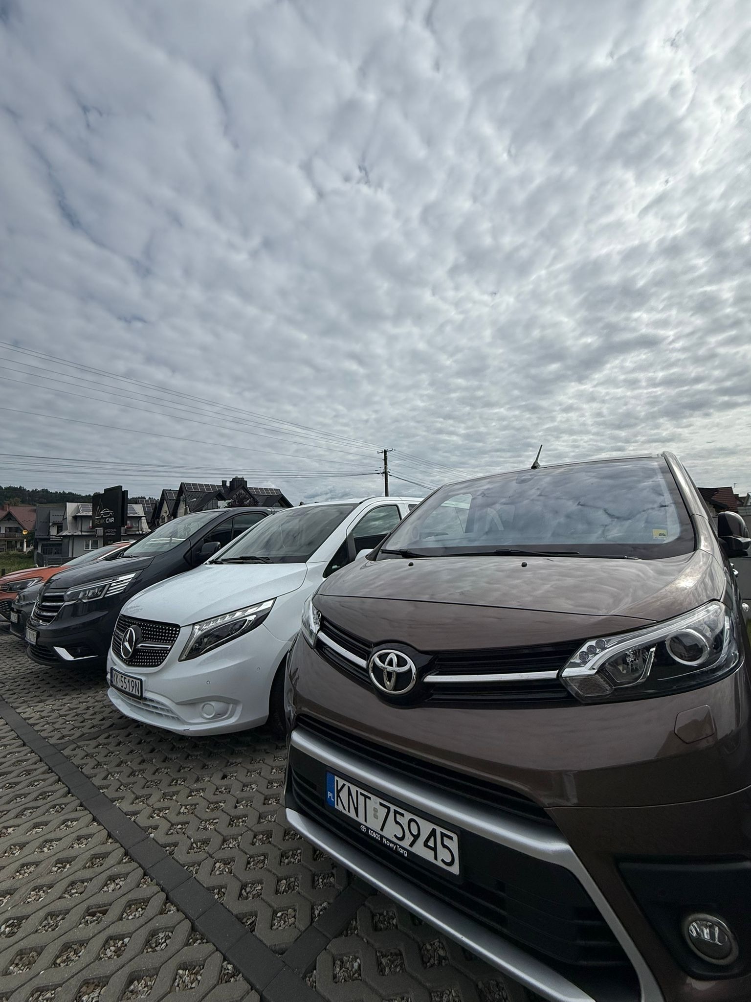 Flota samochodów: brązowa Toyota z przodu, za nią biały Mercedes Vito i czarny Mercedes. Pochmurne niebo w tle, kostka brukowa na parkingu.