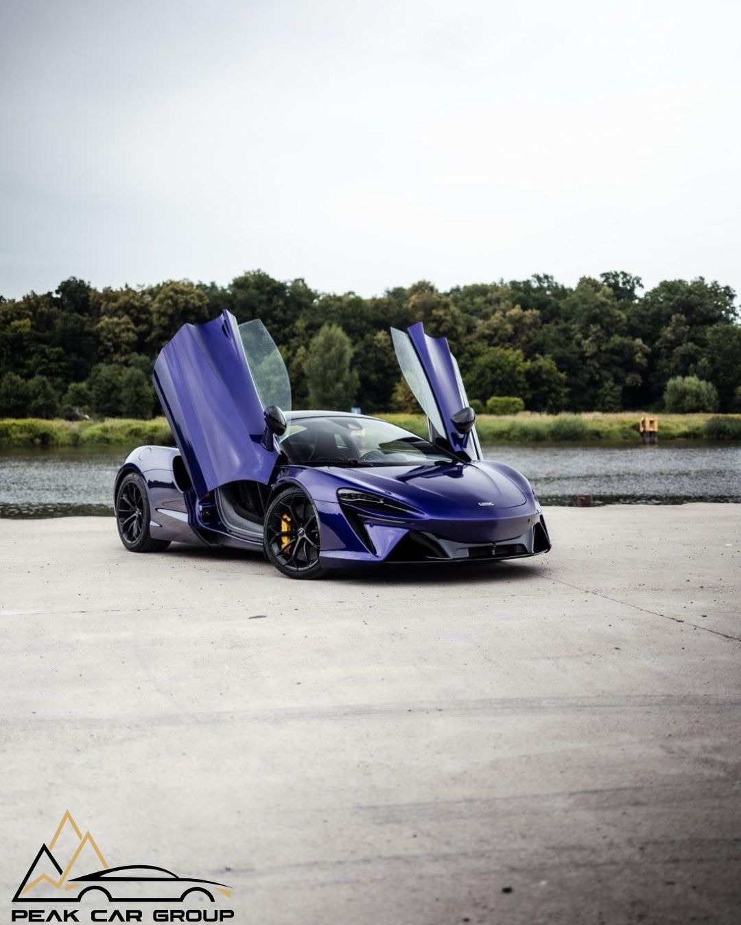 Fioletowy McLaren z otwartymi drzwiami typu 'butterfly' na tle jeziora i drzew. Widoczny logotyp Peak Car Group.