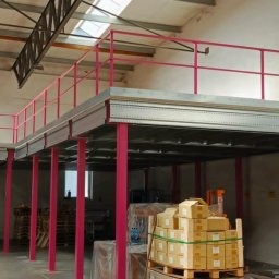 Scr Konstrukcje Piotr Oleszczuk - Wnętrze hali z antresolą o konstrukcji stalowej, pomalowanej na różowo, z widocznymi schodami i paletami z kartonami. Oświetlenie naturalne z okien dachowych.