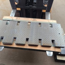 Scr Konstrukcje Piotr Oleszczuk - Stalowe płyty z wycięciami, ułożone na drewnianych belkach, transportowane wózkiem widłowym. Widoczna faktura blachy ryflowanej i elementy konstrukcyjne wózka.