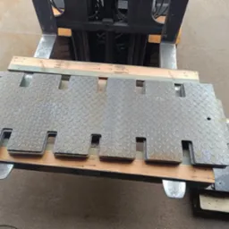 Stalowe płyty z wycięciami, ułożone na drewnianych belkach, transportowane wózkiem widłowym. Widoczna faktura blachy ryflowanej i elementy konstrukcyjne wózka.