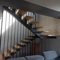 Nowoczesne schody drewniane z metalową balustradą i konstrukcją, z widocznym fragmentem ceglanej ściany w tle,  w minimalistycznym wnętrzu.