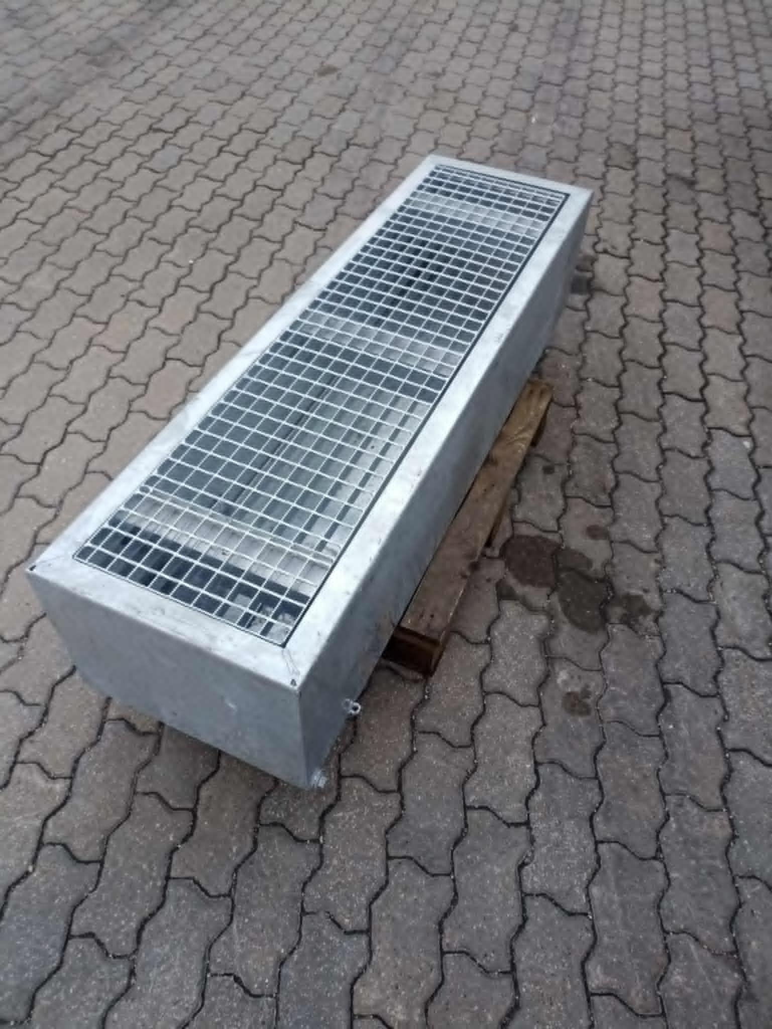 Gotowa, metalowa kratka ściekowa o prostokątnym kształcie, umieszczona na drewnianej palecie, na tle szarej kostki brukowej.