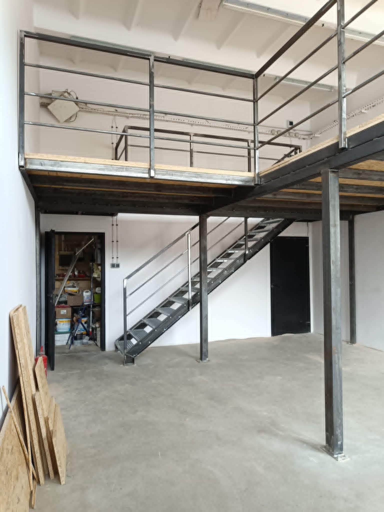 Metalowa konstrukcja antresoli z schodami w surowym wnętrzu. Widoczne elementy drewniane i metalowe. Minimalistyczny design, industrialny styl. Otwarta przestrzeń.