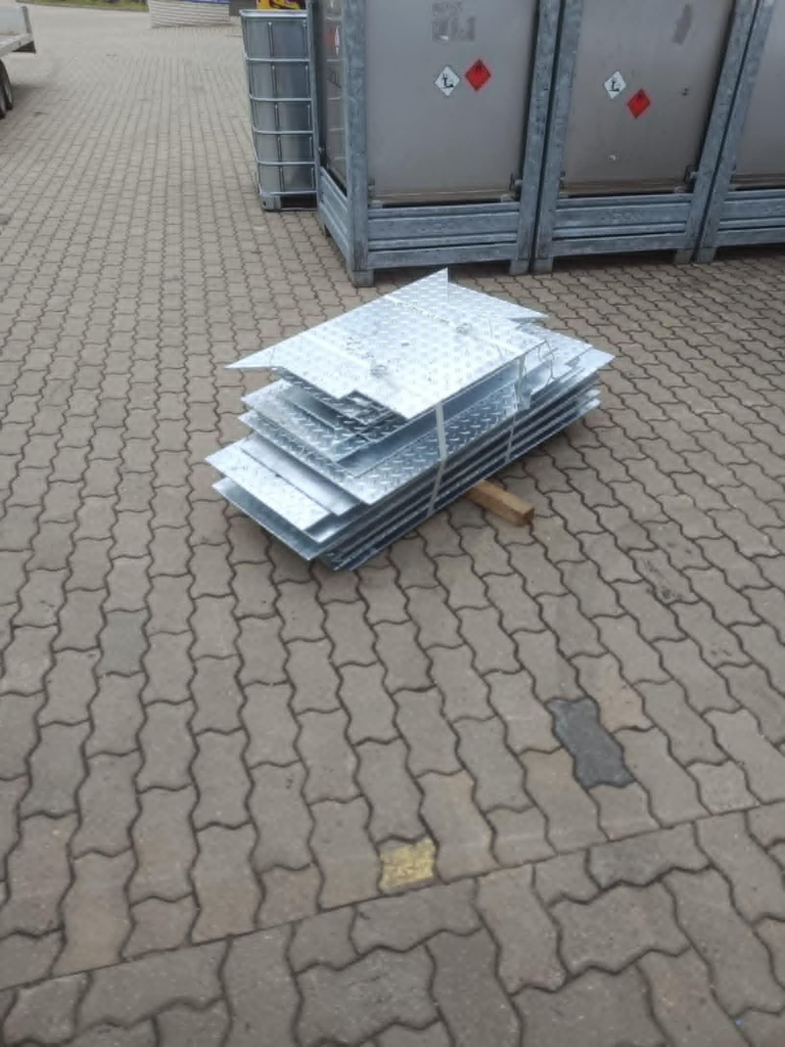 Stalowe, ocynkowane płyty podestowe ułożone w stos na kostce brukowej, zabezpieczone drewnianym klinem. W tle kontenery z oznaczeniami ADR.