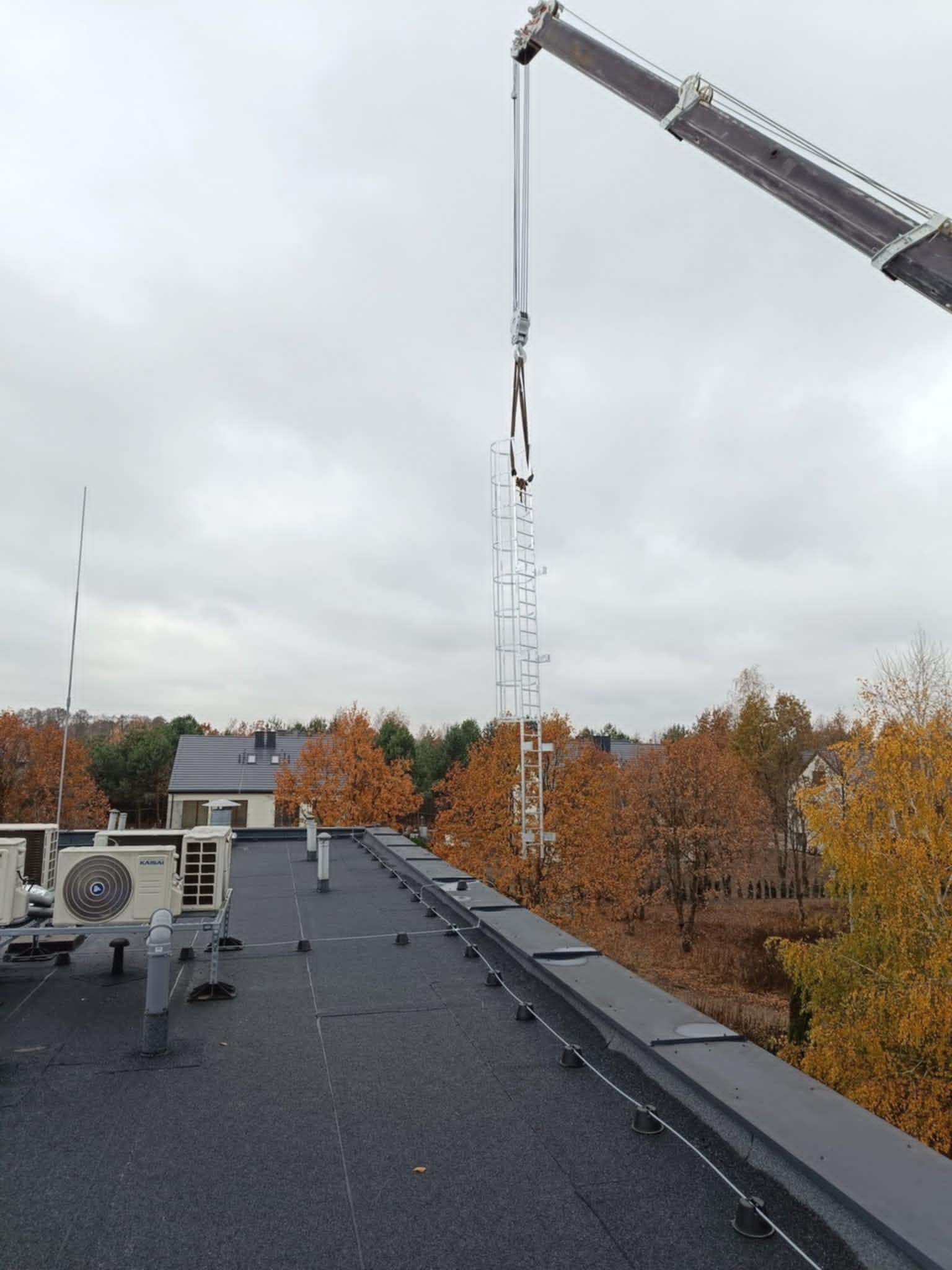 Montaż drabiny ewakuacyjnej na dachu budynku z użyciem dźwigu. Widoczne klimatyzatory i jesienne drzewa w tle. Pochmurne niebo. Kąt ujęcia z dołu.