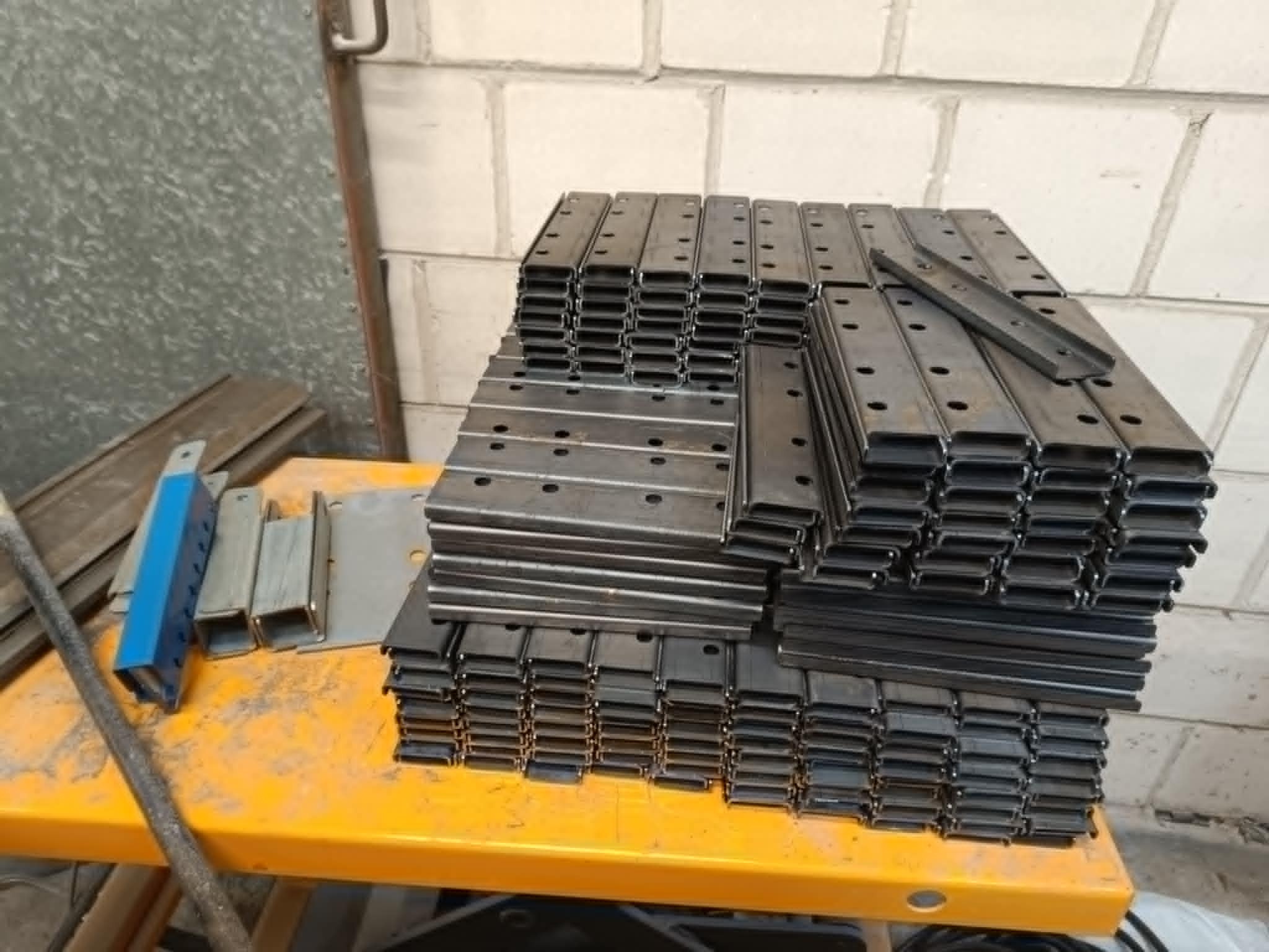 Stos metalowych profili C z otworami, leżących na żółtym stole warsztatowym, na tle ściany z białej cegły. Widoczne fragmenty innych metalowych elementów.