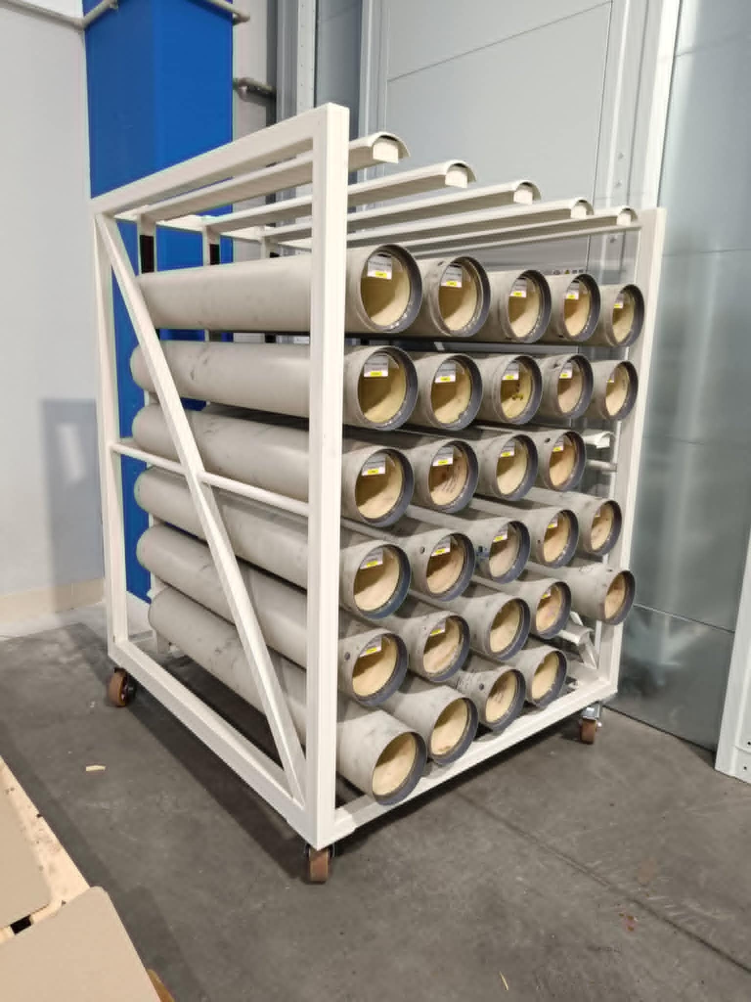 Wózek magazynowy z rurami o szarym odcieniu, każda z etykietą. Metalowa konstrukcja wózka na kółkach. Tło stanowią ściany hali produkcyjnej.