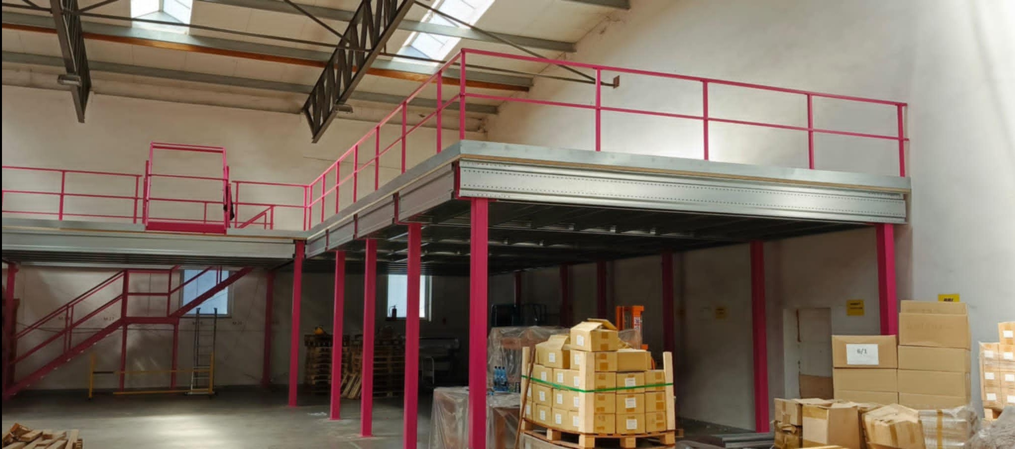 Wnętrze hali z antresolą o konstrukcji stalowej, pomalowanej na różowo, z widocznymi schodami i paletami z kartonami. Oświetlenie naturalne z okien dachowych.