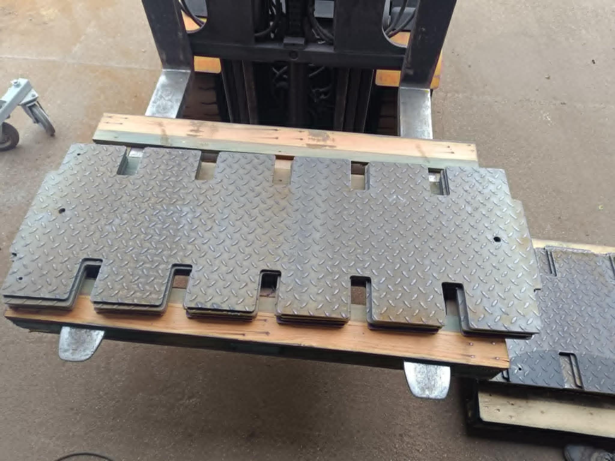 Stalowe płyty z wycięciami, ułożone na drewnianych belkach, transportowane wózkiem widłowym. Widoczna faktura blachy ryflowanej i elementy konstrukcyjne wózka.