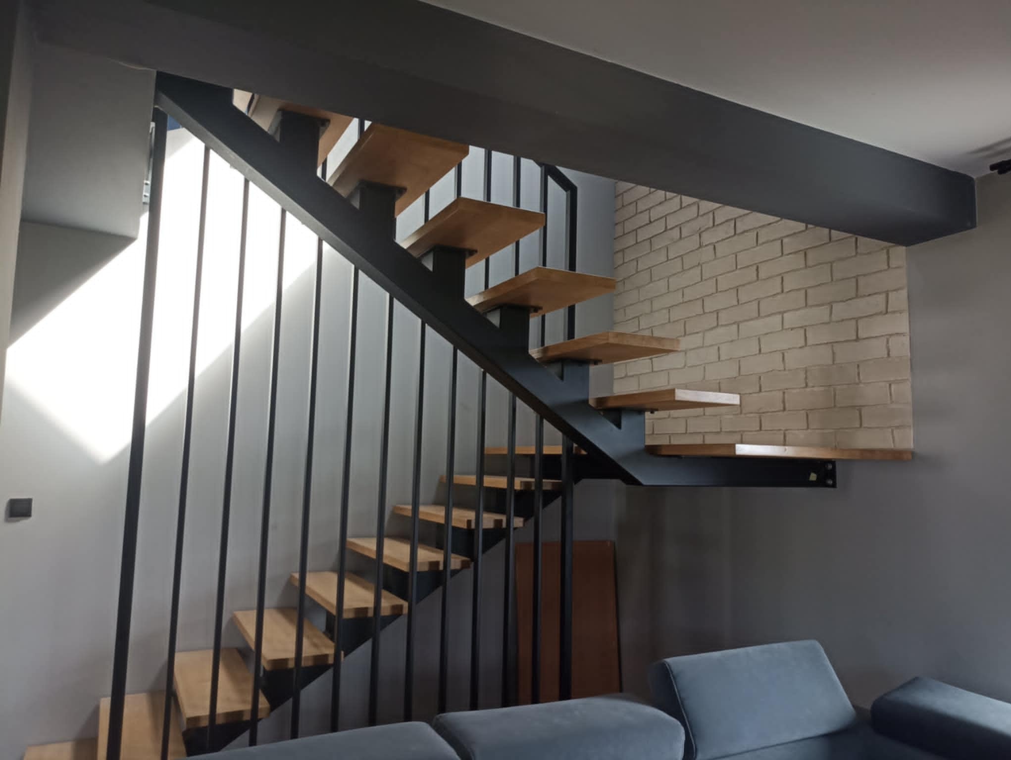 Nowoczesne schody drewniane z metalową balustradą i konstrukcją, z widocznym fragmentem ceglanej ściany w tle,  w minimalistycznym wnętrzu.
