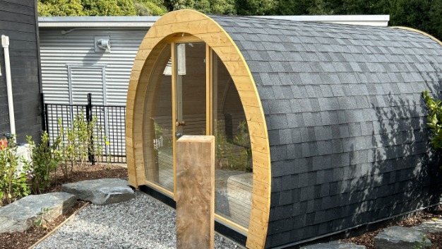 Gotowa, beczkowata sauna ogrodowa z drewnianą ramą i dachem krytym gontem bitumicznym, ustawiona na żwirowym podłożu, w otoczeniu zieleni.