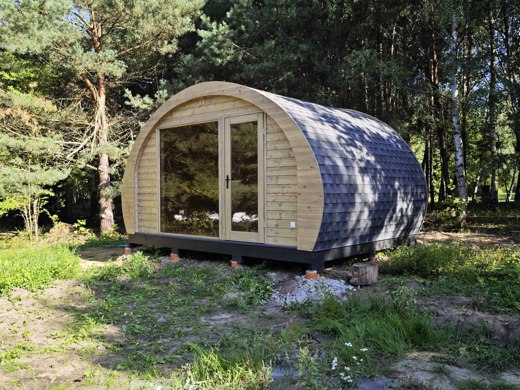 Gotowa, drewniana sauna w kształcie kapsuły, z dwuskrzydłowymi drzwiami, pokryta gontem bitumicznym, ustawiona na podwyższeniu w otoczeniu drzew.