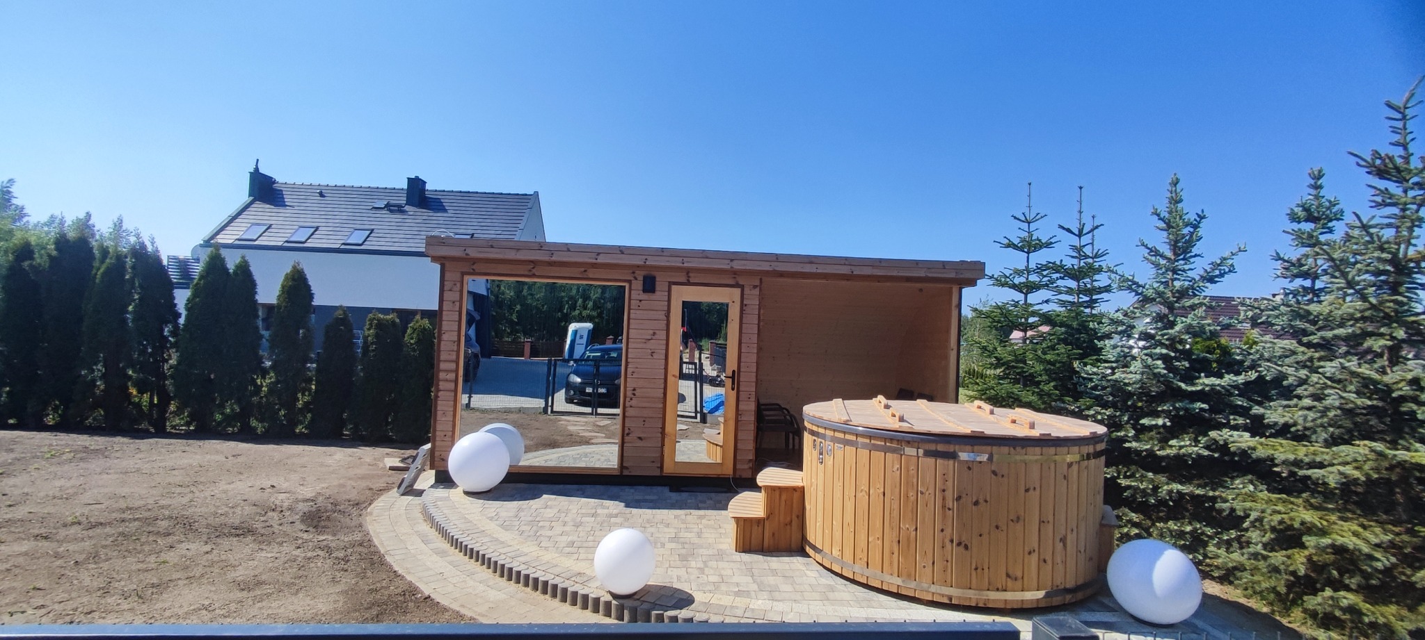 Drewniana sauna z bali i beczka z hydromasażem na zewnątrz, ustawione na brukowanym patio. W tle dom i iglaki, całość skąpana w słonecznym, letnim świetle.