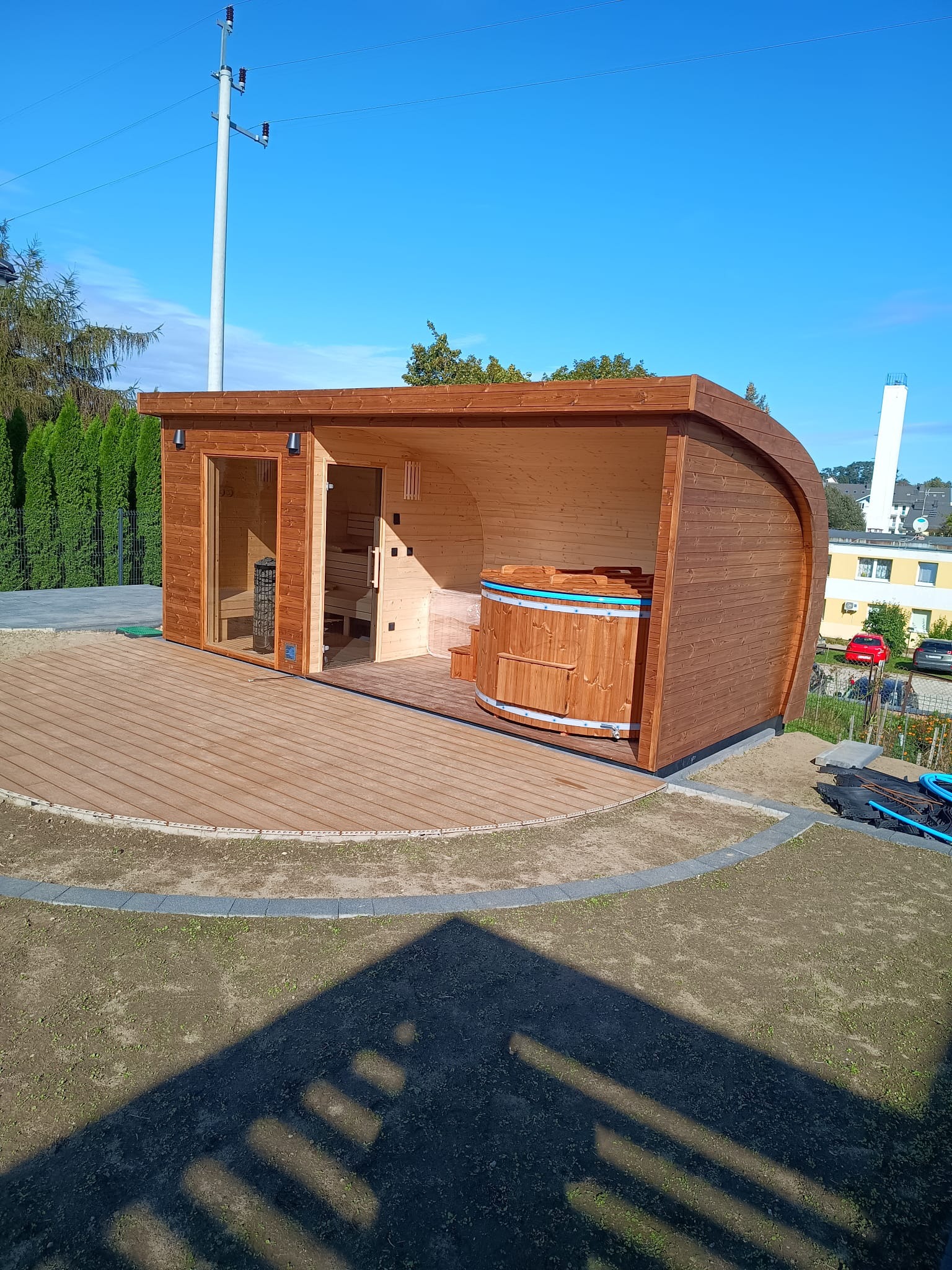 Drewniana sauna beczka z przeszklonym wejściem i jacuzzi na podeście, umiejscowiona w ogrodzie. Elementy architektury i słup elektryczny w tle.