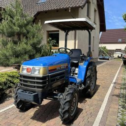 BRONKA GROUND TEAM JAKUB BRONKA - Niebieski traktor SIAL 223 na brukowanej drodze przed domem z zielonymi drzewami. Sprzęt ogrodniczy, gotowy do pracy.