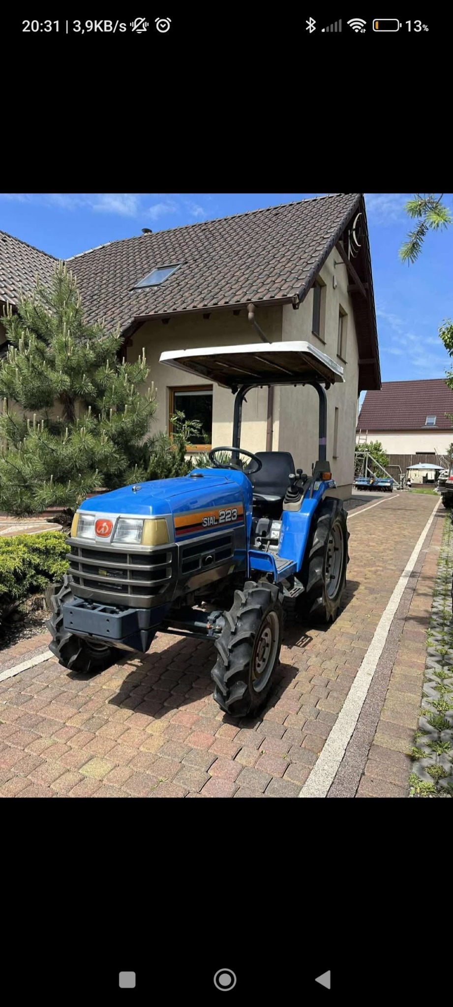 Niebieski traktor SIAL 223 na brukowanej drodze przed domem z zielonymi drzewami. Sprzęt ogrodniczy, gotowy do pracy.