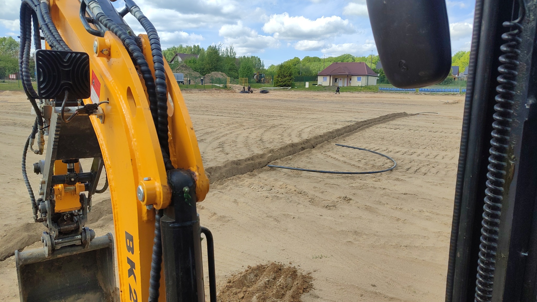 Żółta koparka w Suchorze wykopuje rów pod instalację nawadniającą. Widoczny fragment rury i wyrównany teren. W tle dom i drzewa.