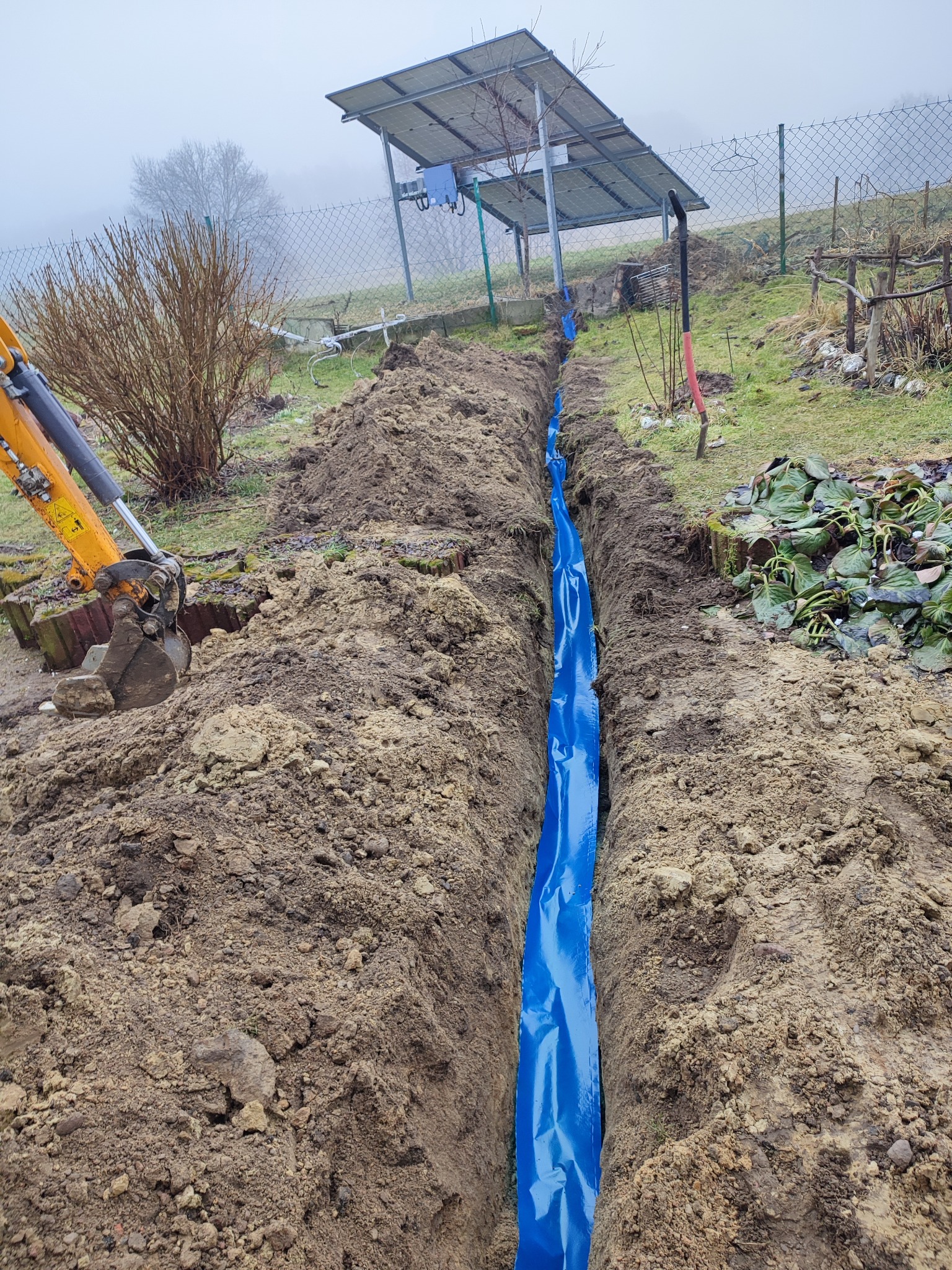 Wykopany rów z niebieską folią, częściowo widoczny mały koparka, ziemia, w tle roślinność i ogrodzenie. Wykop przygotowany pod instalację.