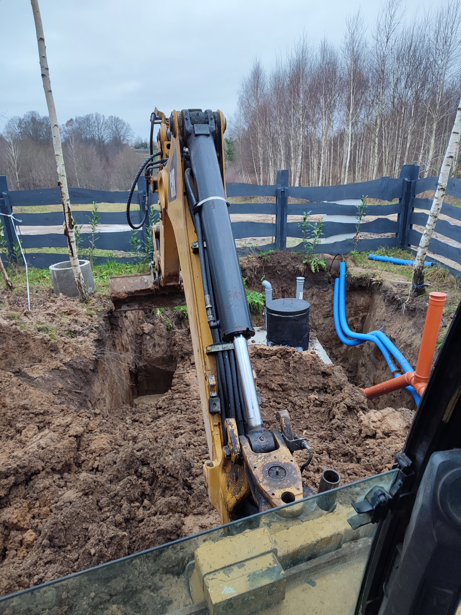 Żółta koparka w trakcie prac ziemnych przy instalacji zbiornika retencyjnego na tle czarnego ogrodzenia i drzew. Widoczne niebieskie rury.