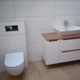 Nowoczesna łazienka z białymi płytkami imitującymi panele, wiszącą toaletą i szafką z umywalką na drewnianym blacie. Minimalistyczny design, jasne wnętrze.