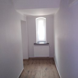 REMONT DOM - Jasne wnętrze po remoncie: drewnopodobna podłoga, białe ściany i sufit, okno z grzejnikiem. Widoczny fragment przejścia do innego pomieszczenia.