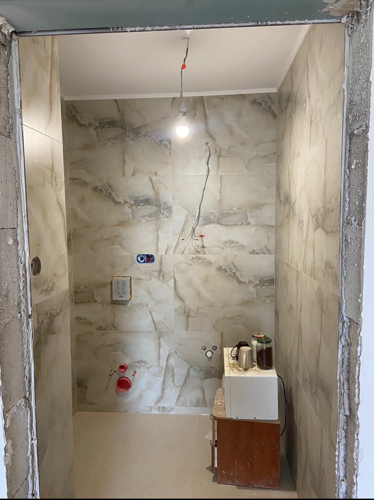 Wykończona łazienka z beżowymi, marmurkowymi płytkami. Widoczne instalacje, gniazdka i niedokończone obramowanie drzwi. Na podłodze płytki w podobnym odcieniu.