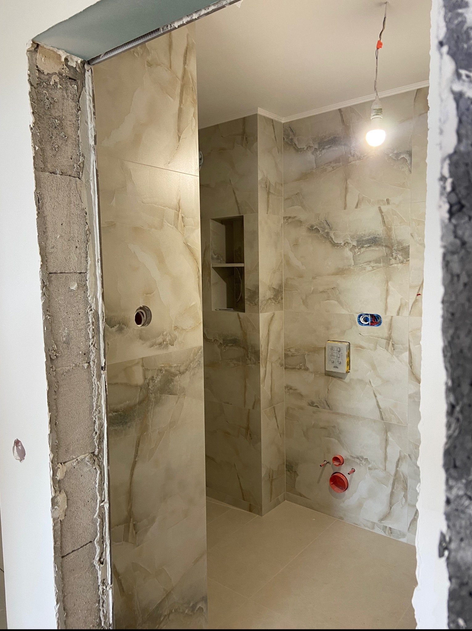 Wykończona łazienka z beżowymi płytkami imitującymi marmur, widoczne fragmenty surowych ścian i instalacji, wnęka z półkami. Ujęcie przez otwór drzwiowy.