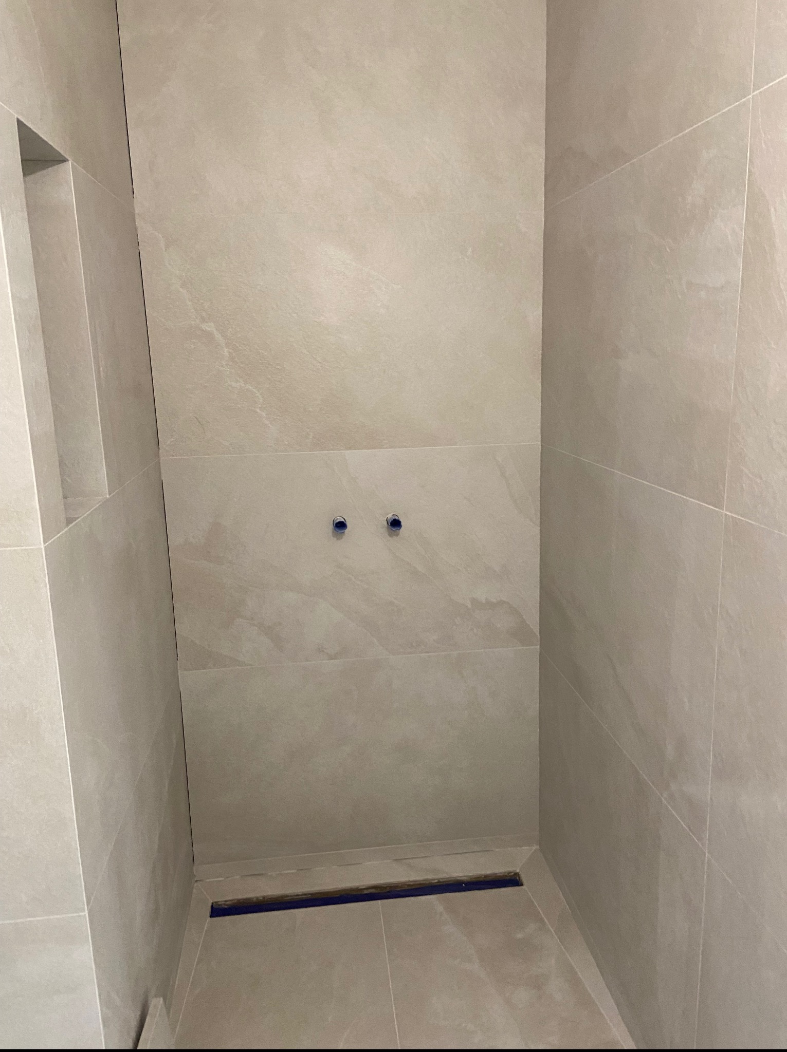 Wykończona, beżowa kabina prysznicowa z odpływem liniowym i wnęką w ścianie. Widoczne dwa wyjścia na baterię. Minimalistyczny styl z delikatnym wzorem na płytkach.