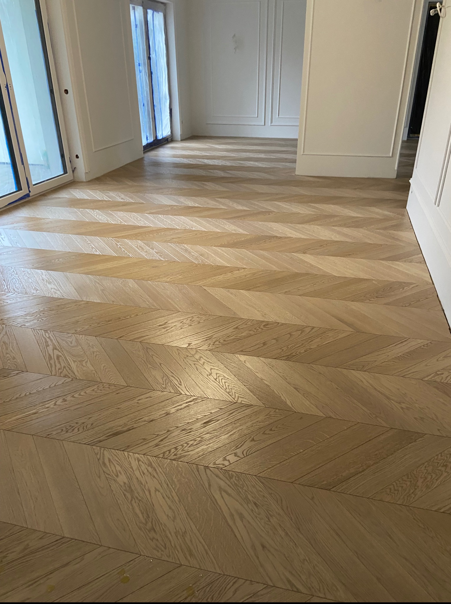 Parkiet ułożony we wzór jodełki francuskiej w jasnym, przestronnym wnętrzu. Widoczne białe listwy przypodłogowe i okna zabezpieczone folią ochronną.
