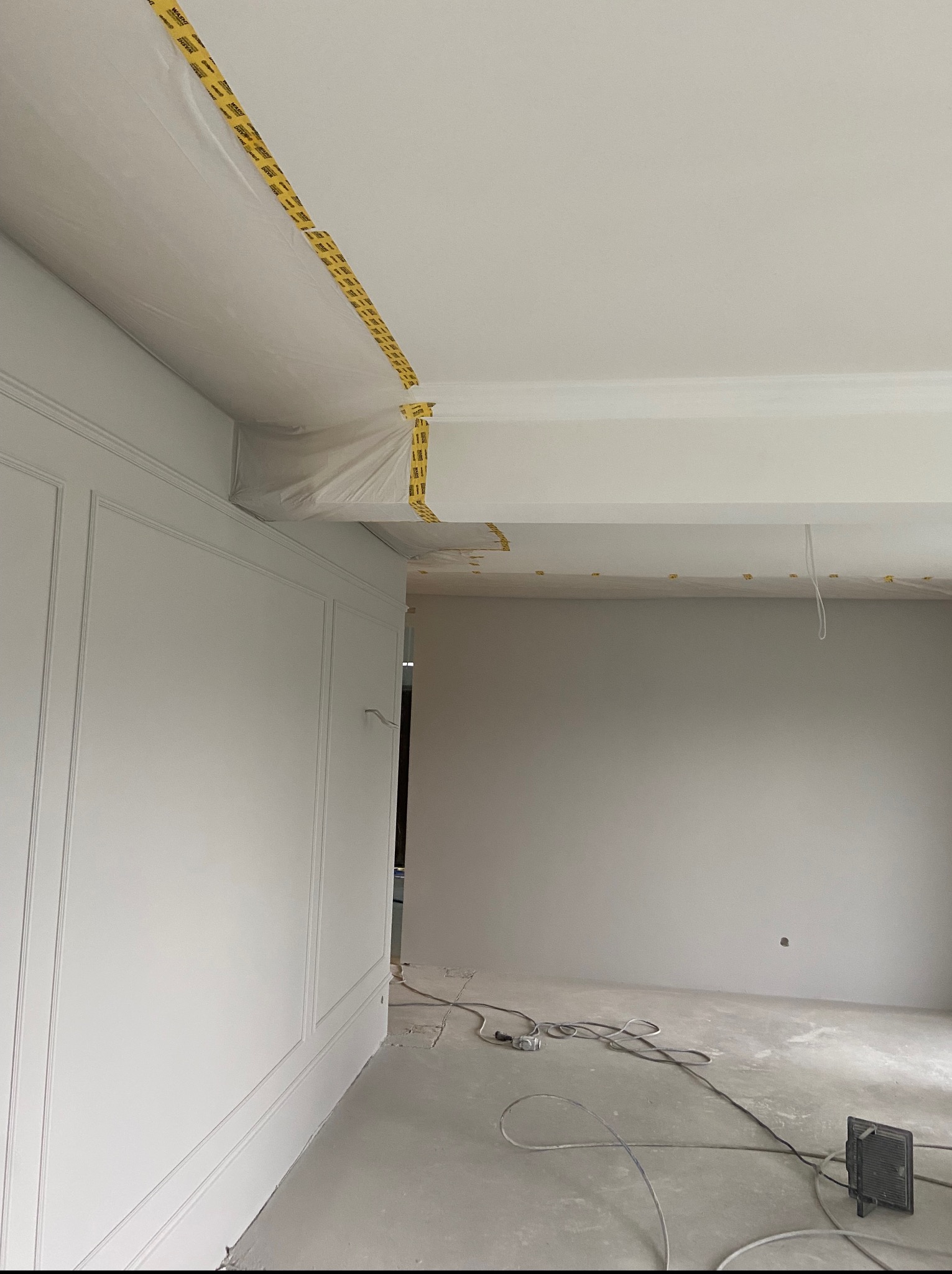 Przygotowanie do malowania: ściany z dekoracyjną sztukaterią zabezpieczone folią i taśmą malarską, surowa podłoga. Widoczne kable i lampa oświetleniowa.