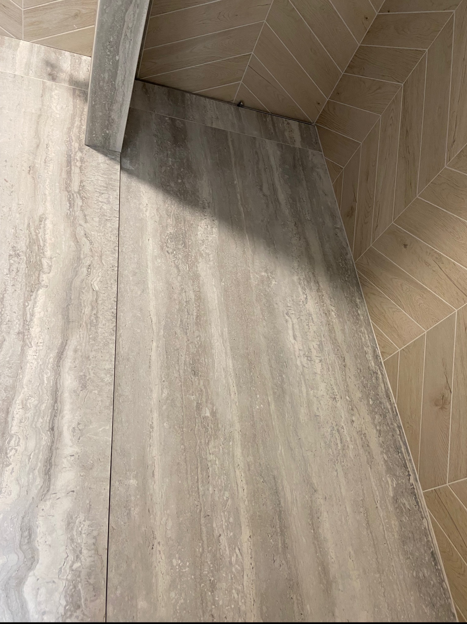 Beżowe płytki imitujące drewno na podłodze i szare płytki z marmurkowym wzorem na ścianie, połączone listwą wykończeniową w łazience. Układ ukośny płytek.