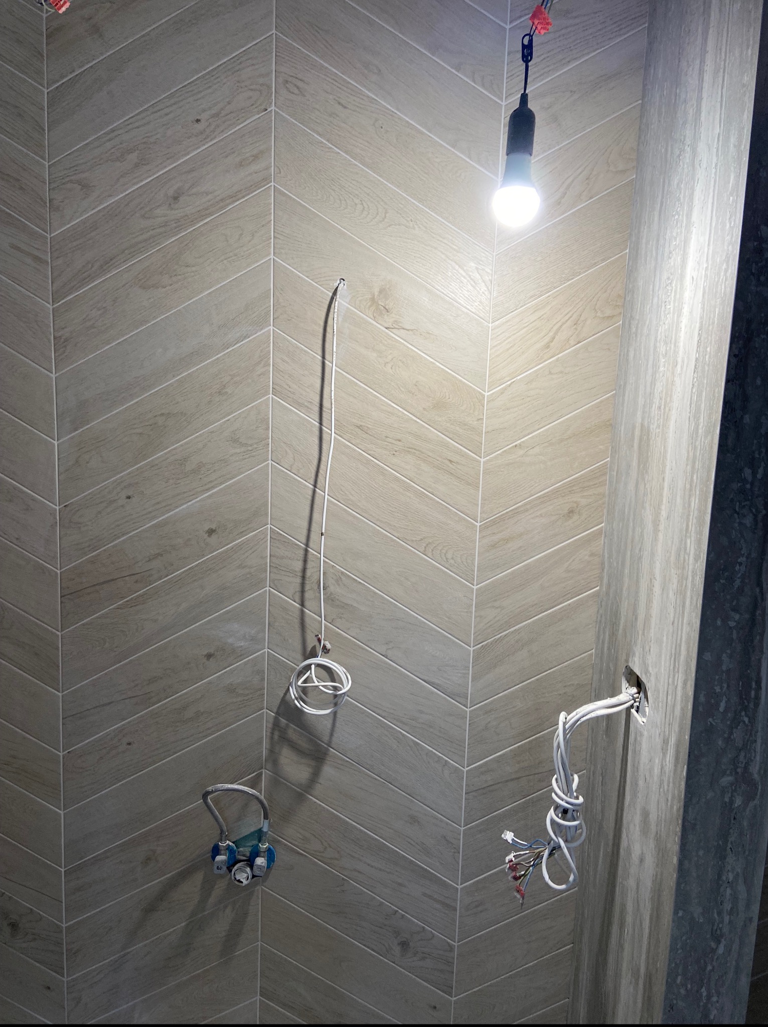 Ściana wyłożona płytkami imitującymi drewno w jodełkę, z widocznymi przewodami i elementami instalacji wodnej w trakcie prac wykończeniowych łazienki.