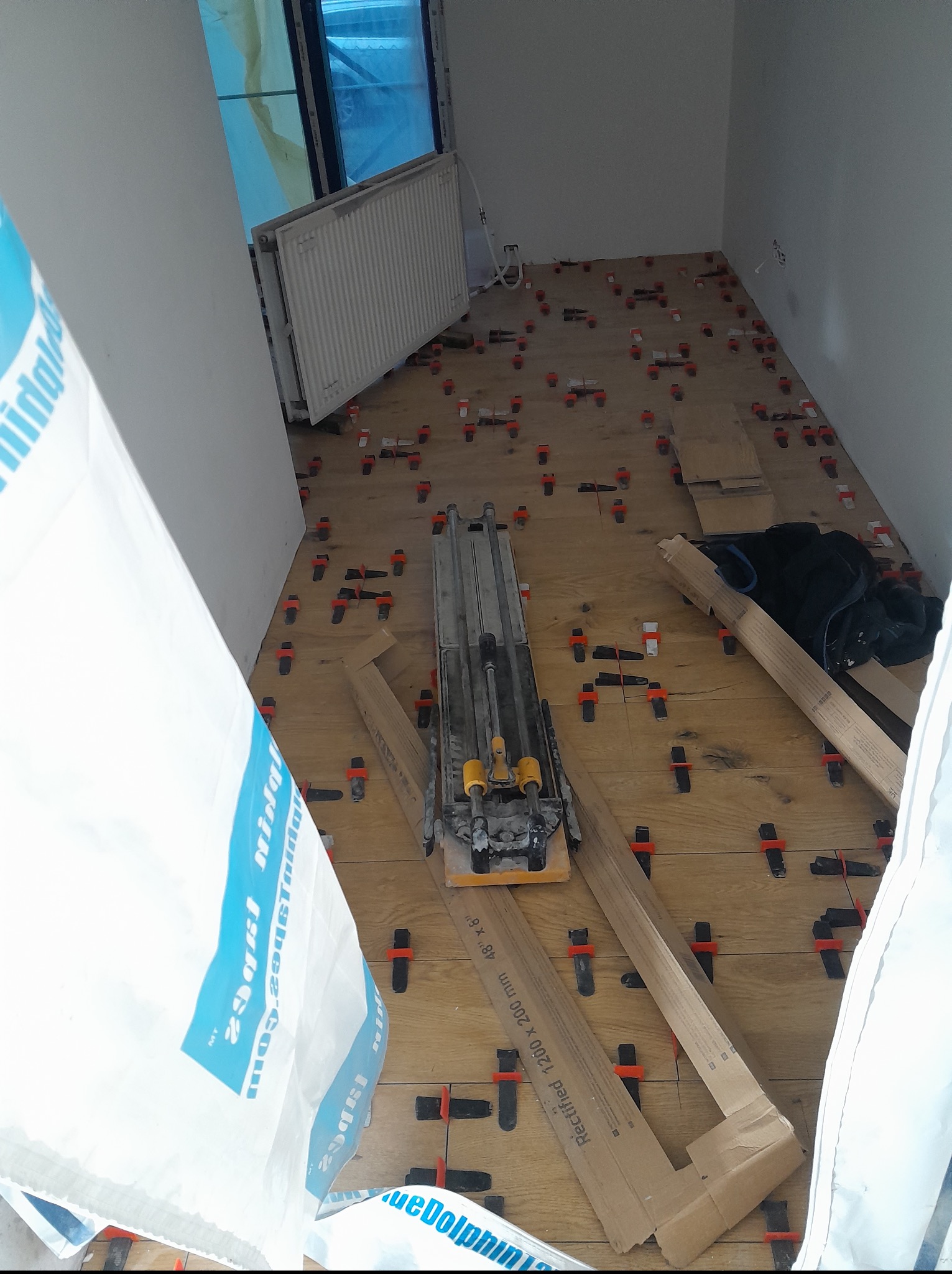 Układanie płytek podłogowych imitujących drewno, z użyciem klipsów poziomujących i narzędzi glazurniczych. Widoczny fragment okna i grzejnika. Praca w toku.