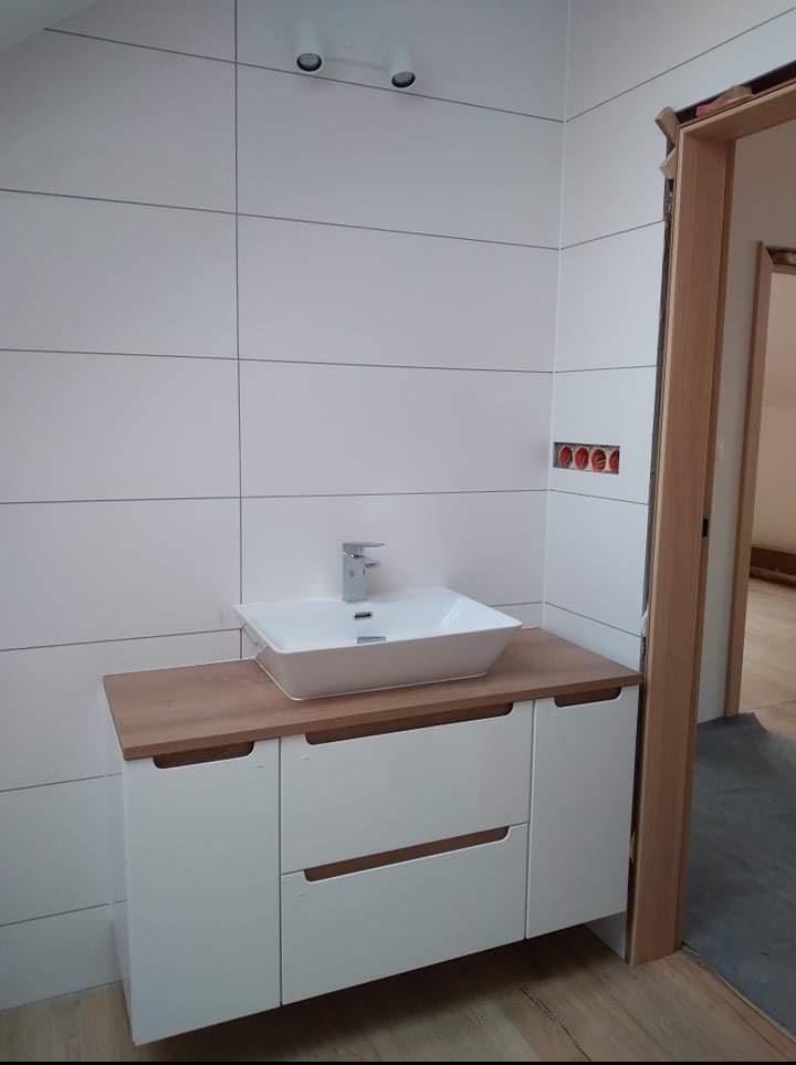 Nowoczesna łazienka z białymi, poziomymi płytkami i szafką z umywalką na drewnianym blacie, widok na ościeżnicę drzwi.