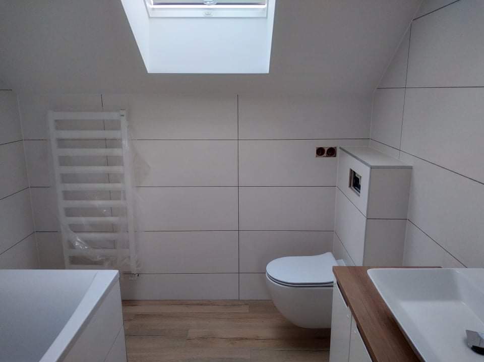 Jasna łazienka na poddaszu z oknem dachowym. Białe, poziome płytki na ścianach, drewnopodobna podłoga, podwieszana toaleta i grzejnik drabinkowy w folii.