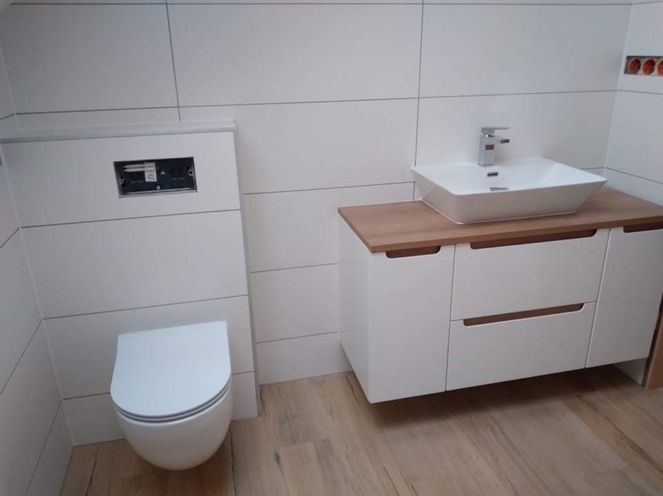 Nowoczesna łazienka z białymi płytkami imitującymi panele, wiszącą toaletą i szafką z umywalką na drewnianym blacie. Minimalistyczny design, jasne wnętrze.