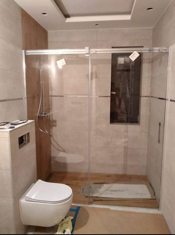 Wykończona łazienka z kabiną prysznicową typu walk-in, beżowe płytki, drewnopodobna ściana, podwieszana toaleta i sufit podwieszany z oświetleniem.