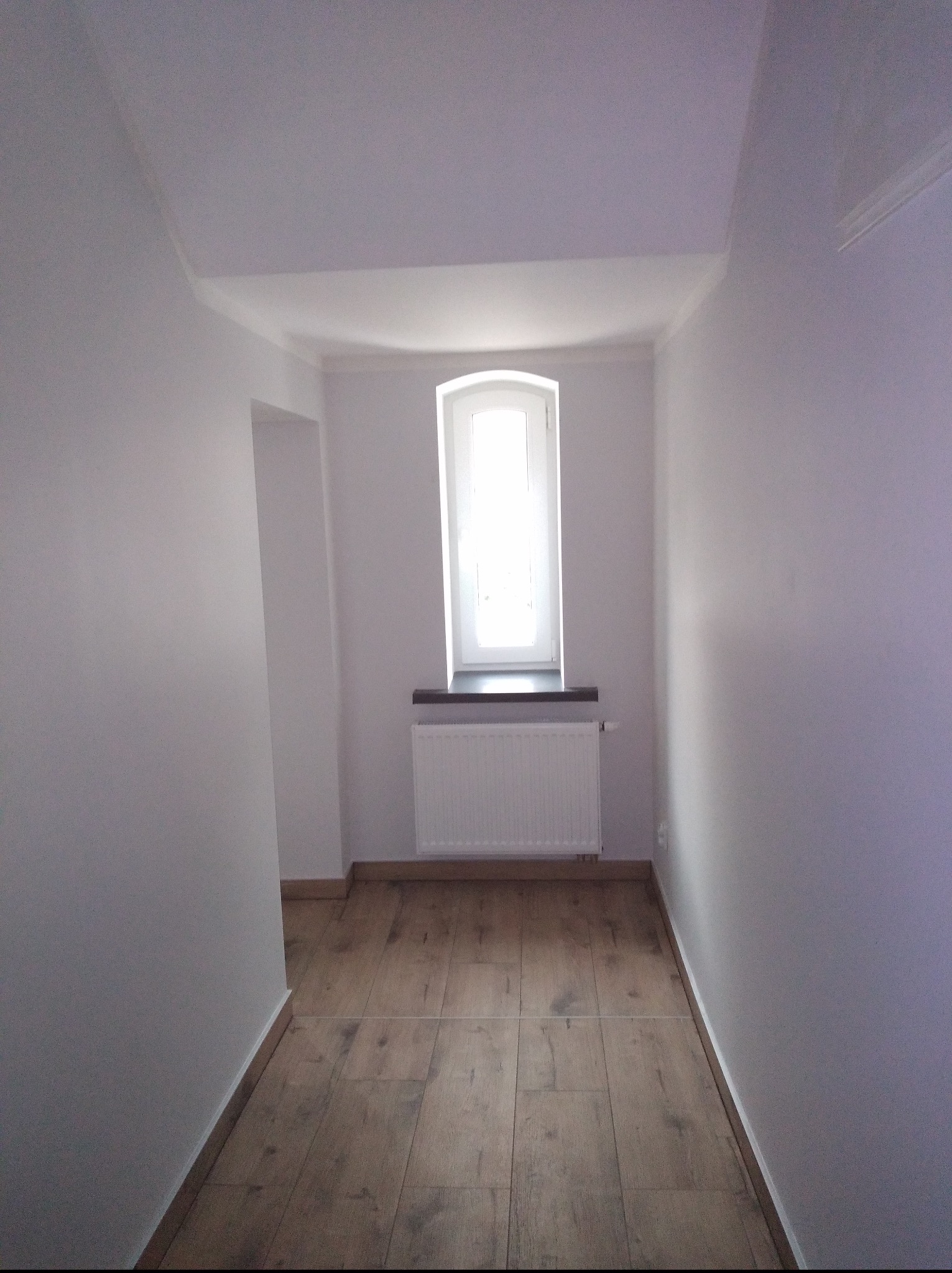 Jasne wnętrze po remoncie: drewnopodobna podłoga, białe ściany i sufit, okno z grzejnikiem. Widoczny fragment przejścia do innego pomieszczenia.