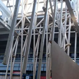 Wnętrze budynku w trakcie budowy z drewnianą konstrukcją szkieletową. Widoczne elementy rusztowania i podnośnika. Oświetlenie dzienne wpada przez okna dachowe.