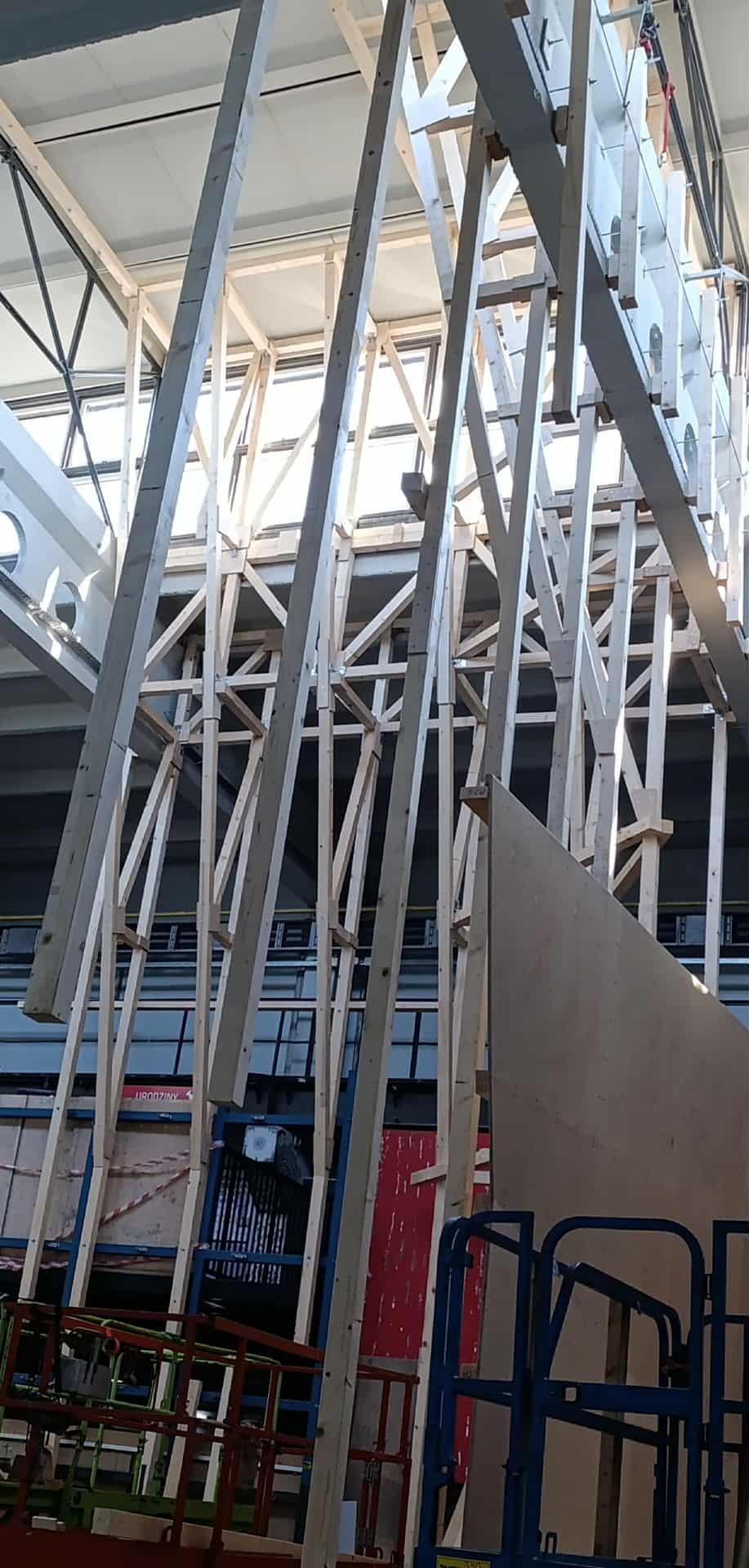 Wnętrze budynku w trakcie budowy z drewnianą konstrukcją szkieletową. Widoczne elementy rusztowania i podnośnika. Oświetlenie dzienne wpada przez okna dachowe.