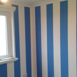Mateusz Trxonkowski - Pomalowane ściany w pionowe, biało-niebieskie pasy w pokoju z oknem. Widoczna listwa przypodłogowa i zabezpieczona podłoga.