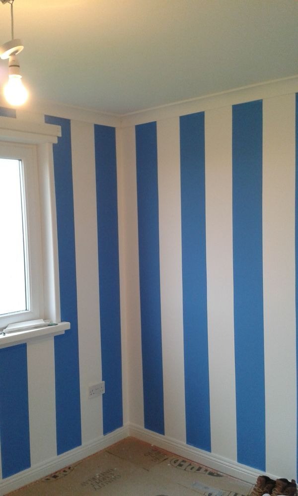 Pomalowane ściany w pionowe, biało-niebieskie pasy w pokoju z oknem. Widoczna listwa przypodłogowa i zabezpieczona podłoga.