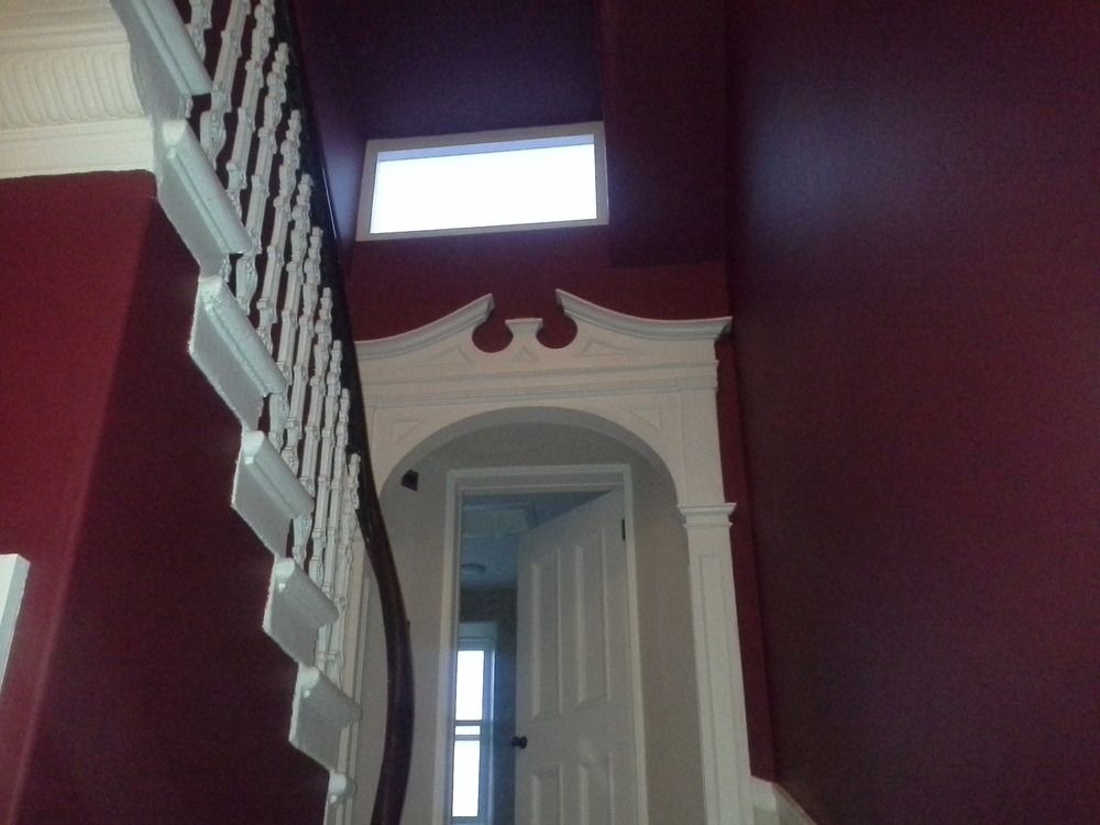 Wnętrze klatki schodowej z bordowymi ścianami, białymi schodami i ościeżnicą. Widoczne drzwi i okno w górnej części ściany. Dekoracyjne elementy architektoniczne.