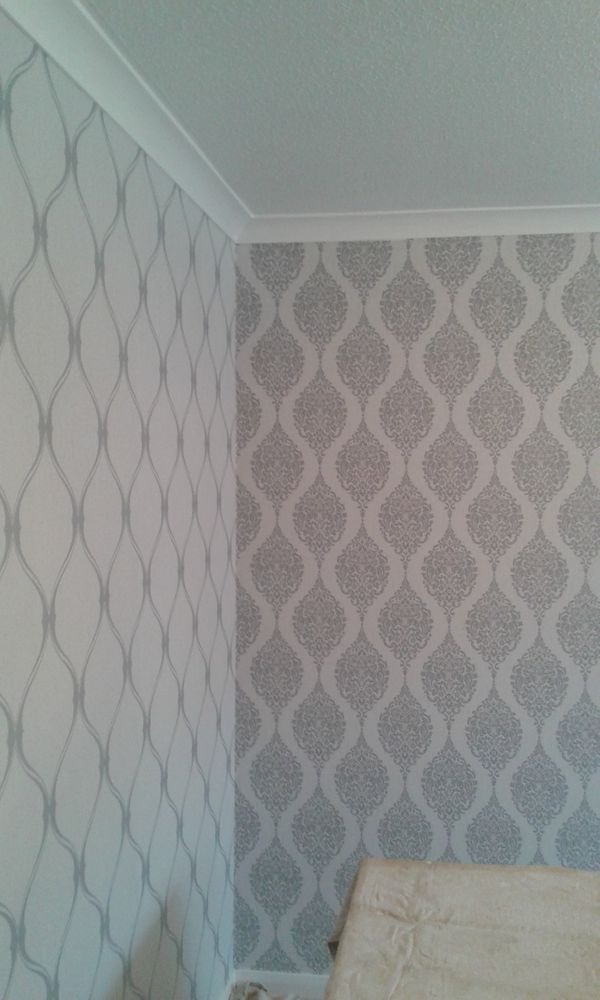 Pokój po remoncie: Dwie ściany z różnymi wzorami tapet w odcieniach szarości, listwa sufitowa. Fragment łóżka w dolnej części kadru.
