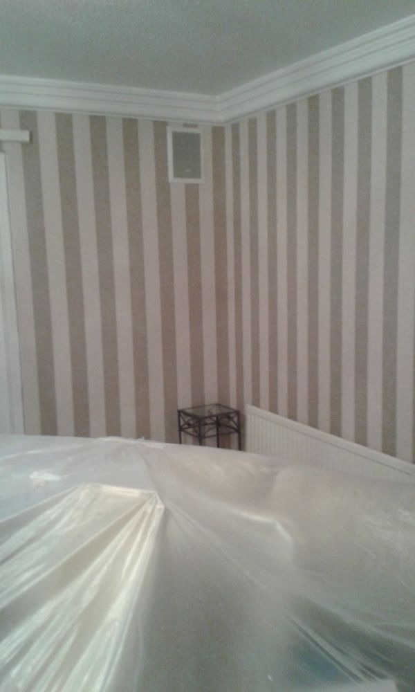 Ściana w pionowe pasy beżu i bieli, zabezpieczona folią malarską. Widoczny fragment grzejnika, stolika i głośnika w ścianie. Listwa dekoracyjna pod sufitem.